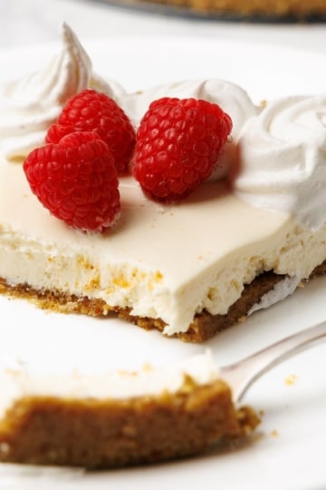 A slice of no-bake cheesecake on a white plate with whipped cream, raspberries, and a fork with a bite taken