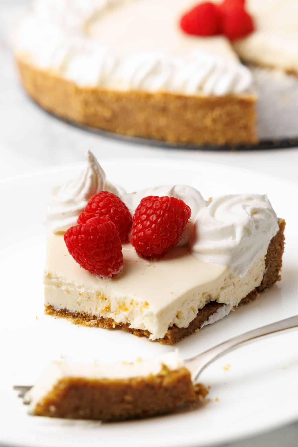 A slice of no-bake cheesecake with whipped cream rosettes and raspberries, golden almond flour crust visible, whole cheesecake in the background
