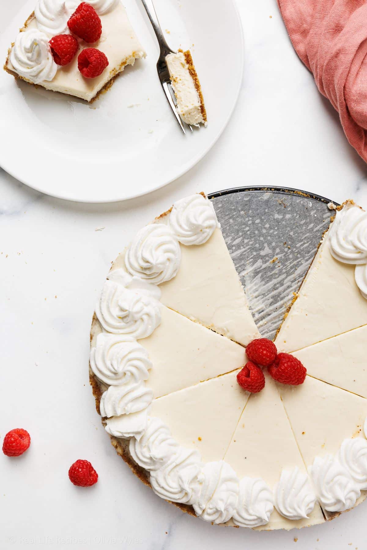 Overhead view of a sliced no-bake cheesecake with whipped cream rosettes and raspberries, one slice on a plate with a fork