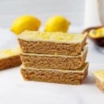 Stack of three lemon protein bars with a white chocolate top dusted with lemon zest, two whole lemons softly blurred in the background.