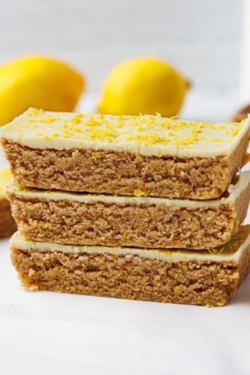 Stack of three lemon protein bars with a white chocolate top dusted with lemon zest, two whole lemons softly blurred in the background.