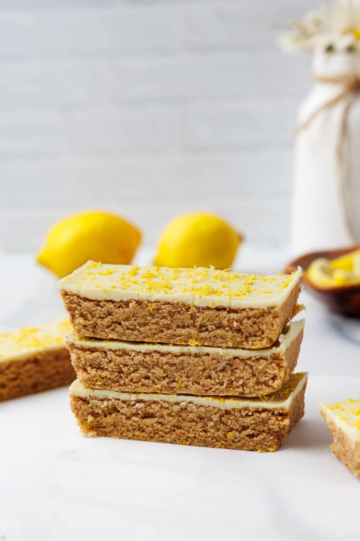 Three lemon protein bars stacked with a white chocolate and lemon zest top, two whole lemons and a small white flower vase softly blurred behind them.