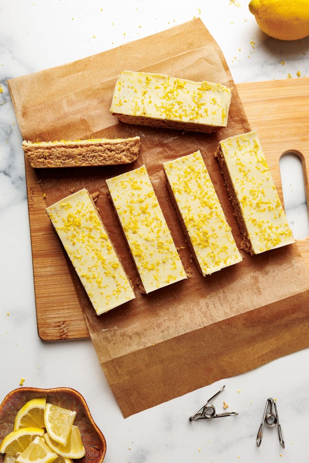 Six sliced lemon protein bars laid on parchment on a wooden cutting board, each topped with set white chocolate and lemon zest.