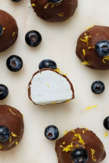 Overhead view of chocolate-coated blueberry lemon truffles on a marble surface, one cut in half to reveal a creamy white filling, scattered with fresh blueberries and lemon zest