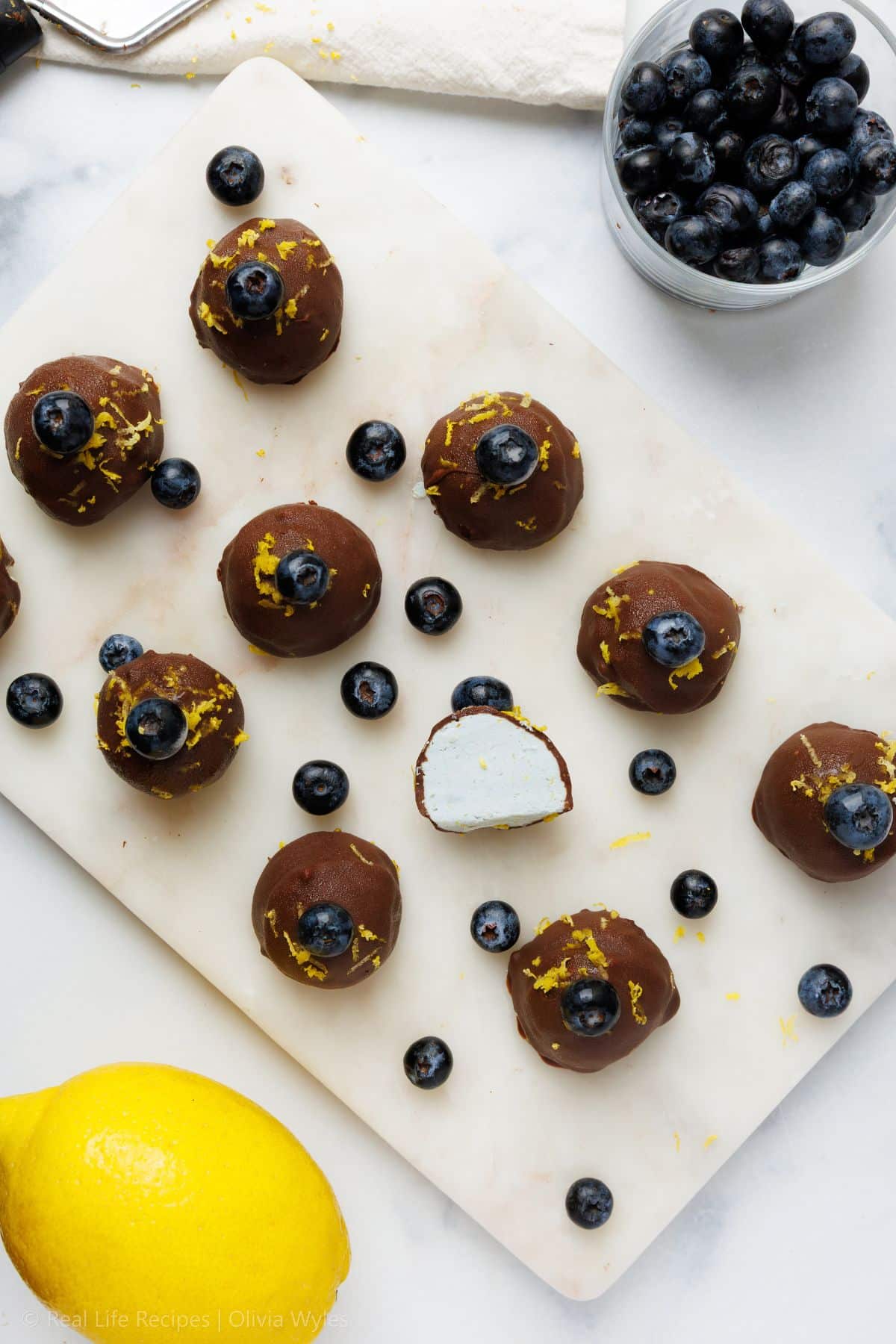 Overhead view of chocolate blueberry truffles on a marble board, one broken open to show a white cream cheese center, scattered with fresh blueberries and a lemon