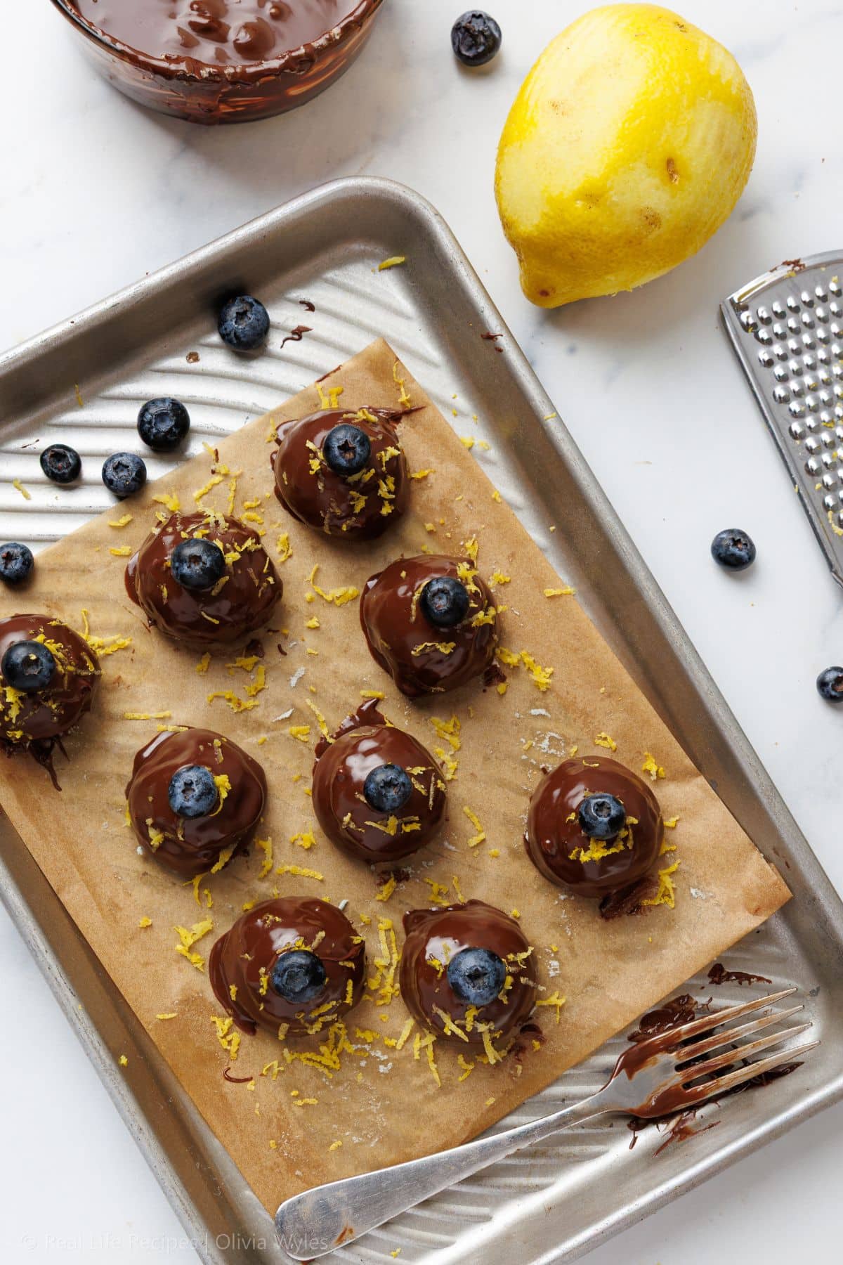 Chocolate-dipped blueberry truffles on a parchment-lined baking sheet, topped with fresh blueberries and lemon zest, with a lemon and grater alongside
