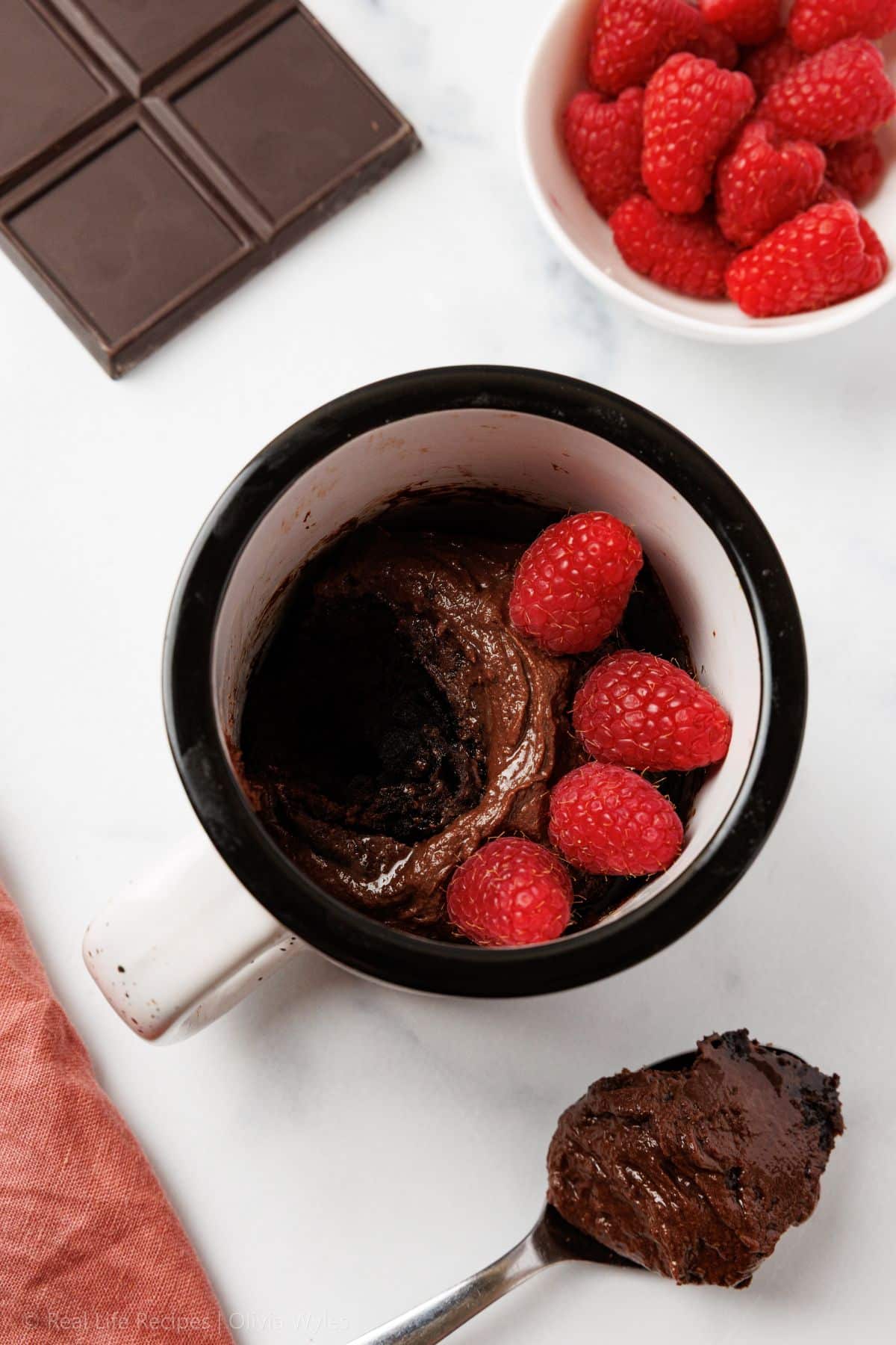 Slightly angled overhead view of a chocolate mug cake topped with raspberries in a black and white mug, with a chocolate-coated spoon resting beside it