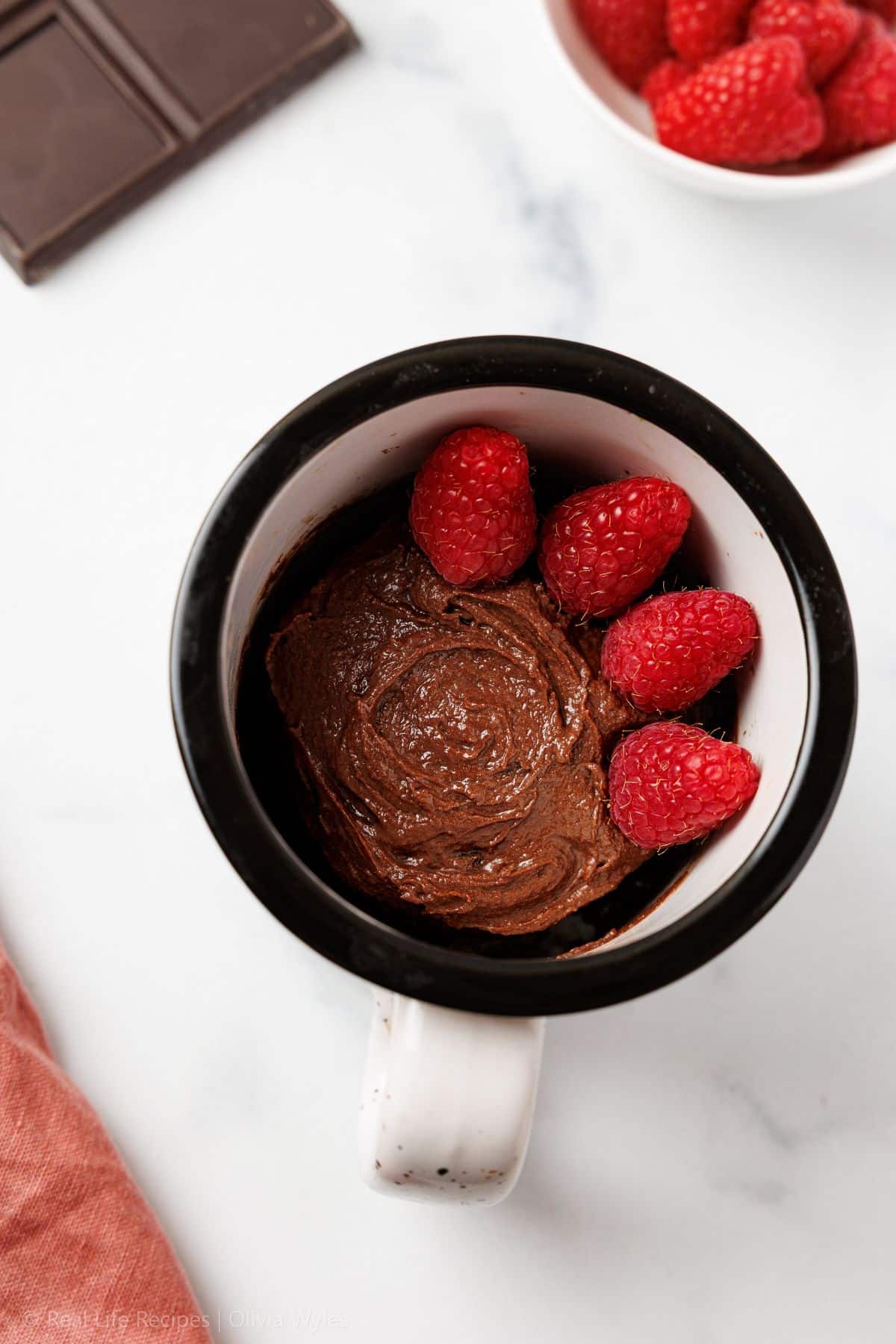 Overhead view of a chocolate mug cake in a black and white ceramic mug, topped with fresh raspberries, on a marble surface with a chocolate bar and bowl of raspberries in the background.