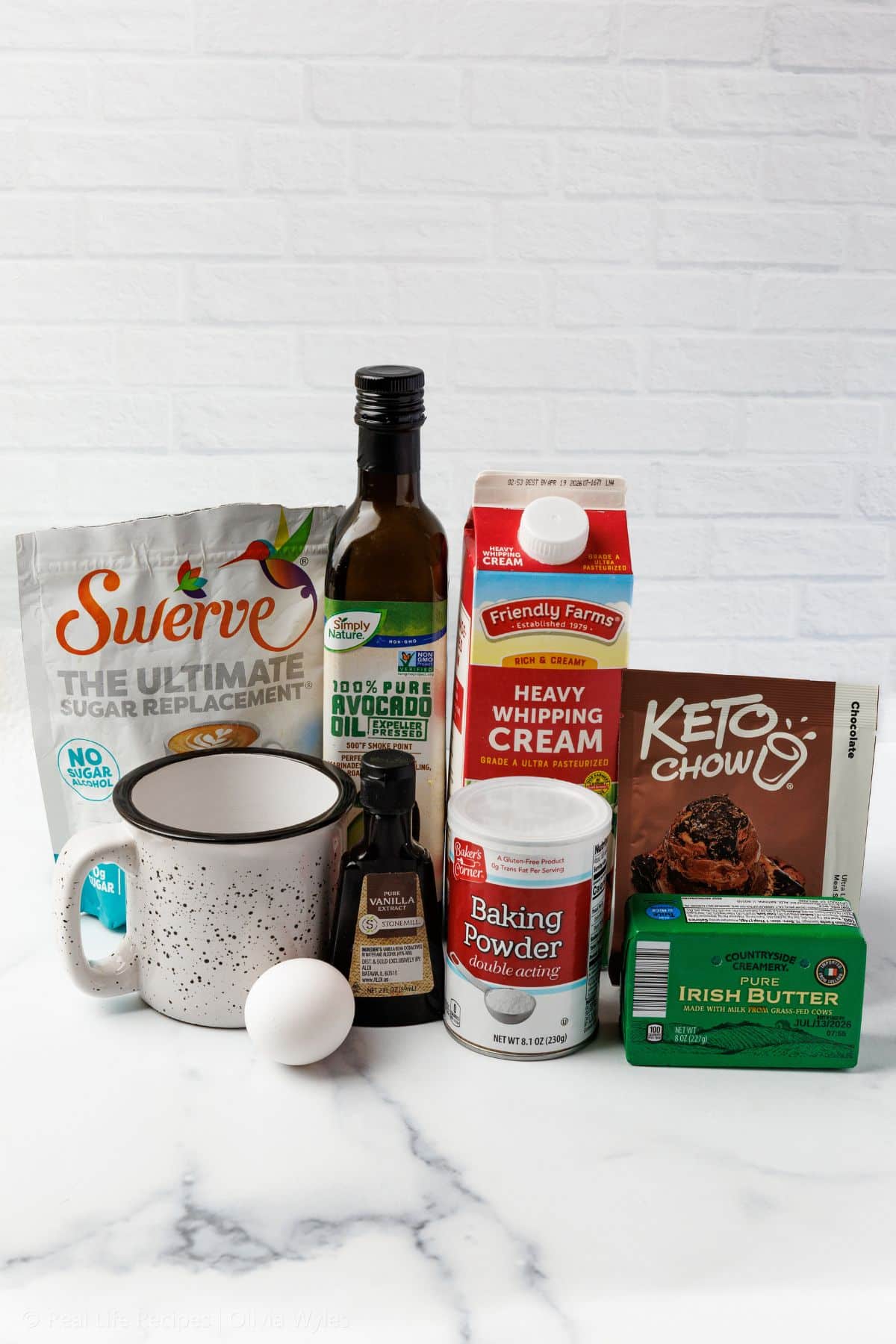 Keto chocolate mug cake ingredients arranged on a marble surface, including Swerve sweetener, Keto Chow, heavy whipping cream, baking powder, Irish butter, vanilla extract, and an egg, with a speckled ceramic mug