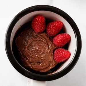 Overhead view of a fudgy keto chocolate mug cake with a swirled top, topped with four fresh raspberries, in a black and white ceramic mug on a white marble surface