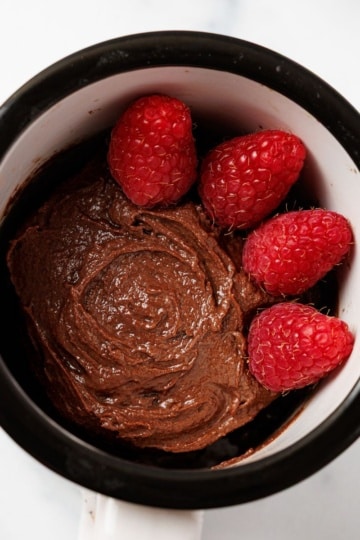 Overhead view of a fudgy keto chocolate mug cake with a swirled top, topped with four fresh raspberries, in a black and white ceramic mug on a white marble surface