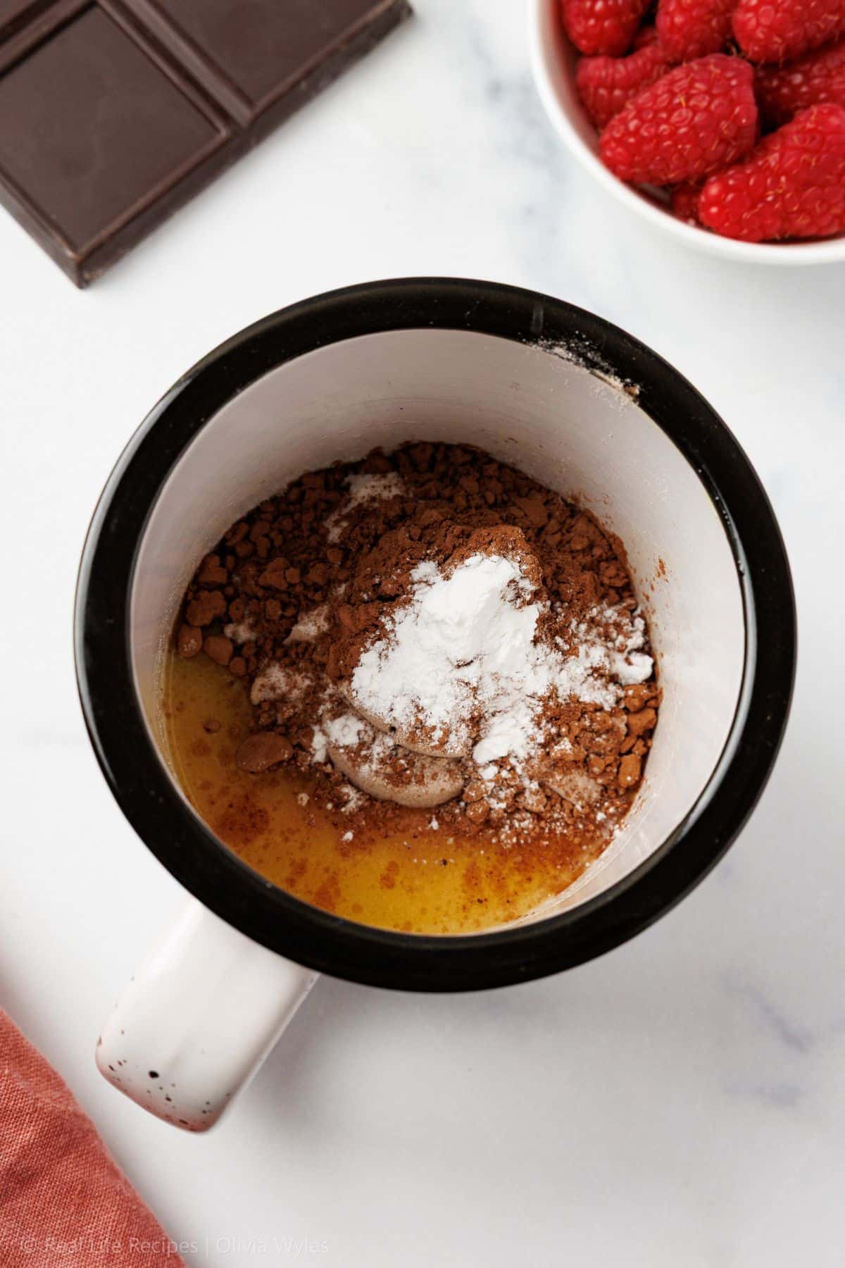 Dry keto mug cake ingredients - cocoa powder, sweetener, and baking powder - added over melted butter and egg in a black and white ceramic mug, ready to mix