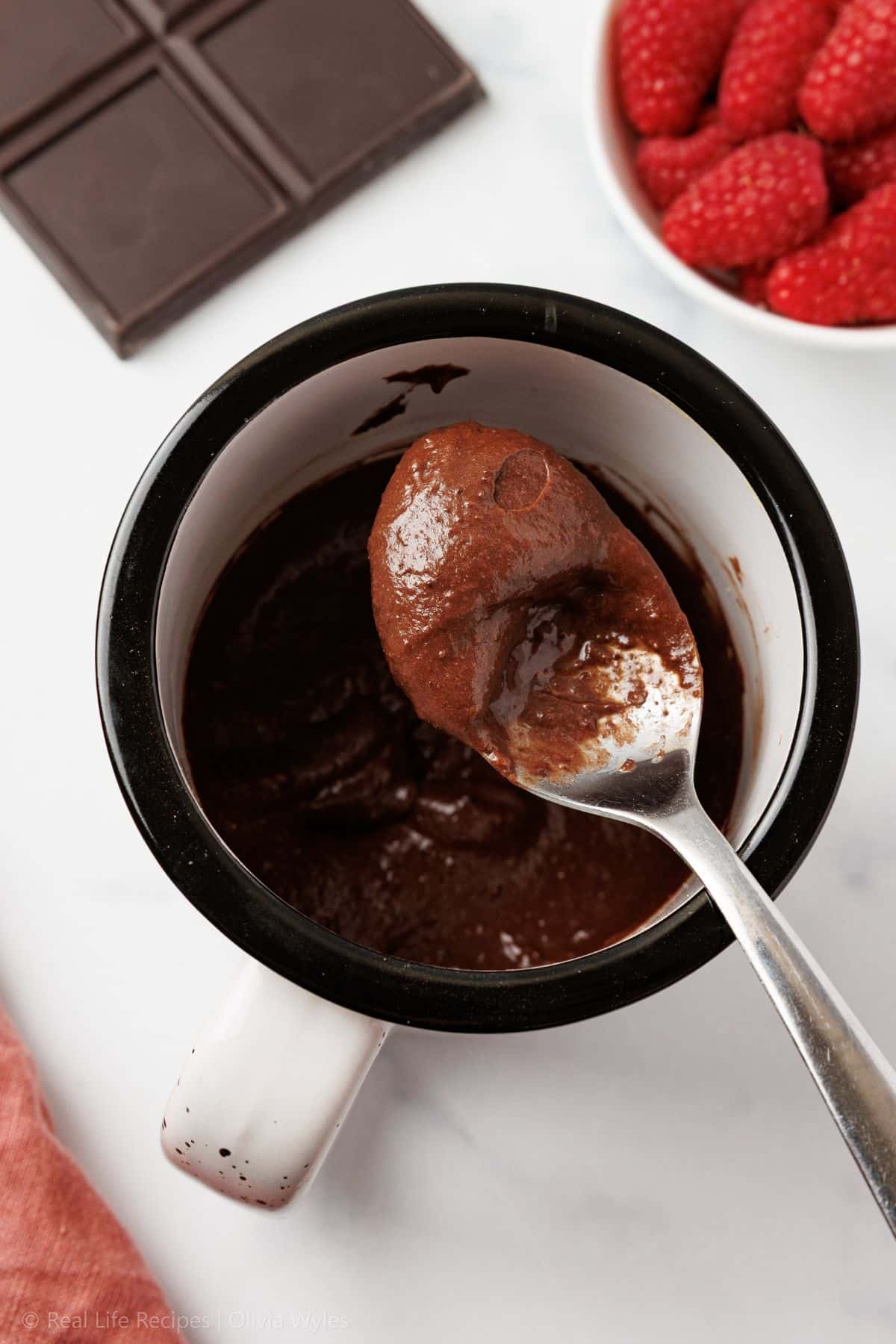 Overhead view of unbaked chocolate mug cake batter being stirred with a spoon in a black and white ceramic mug, with a dark chocolate bar and raspberries in the background