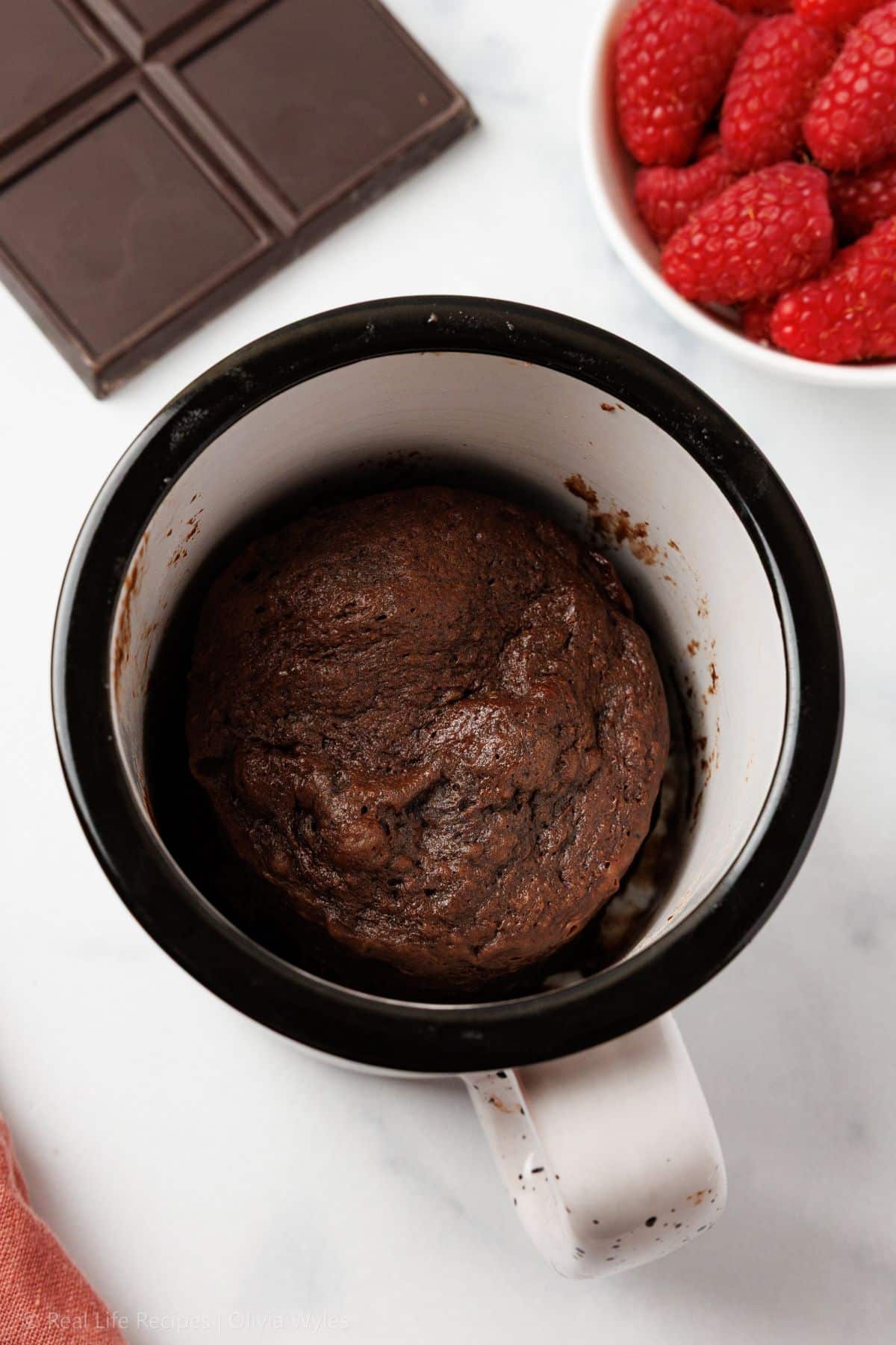 Overhead view of a just-microwaved chocolate mug cake in a black and white ceramic mug, showing a domed, fudgy top before toppings are added