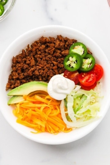 Easy high protein keto taco bowl with ground beef, shredded cheddar, avocado, and fresh toppings in a white bowl.
