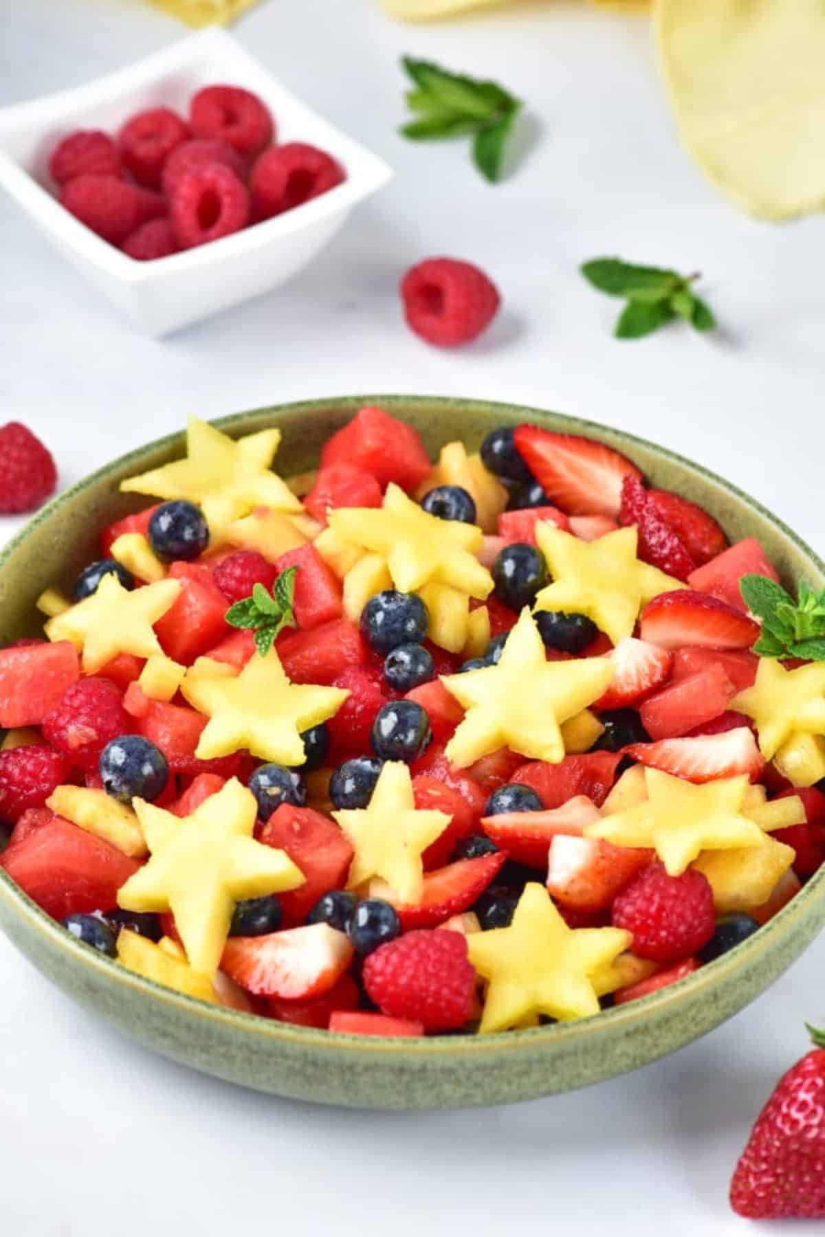 Patriotic fruit salad with fresh strawberries, blueberries, raspberries, and star-shaped pineapple in a green bowl on a white surface.