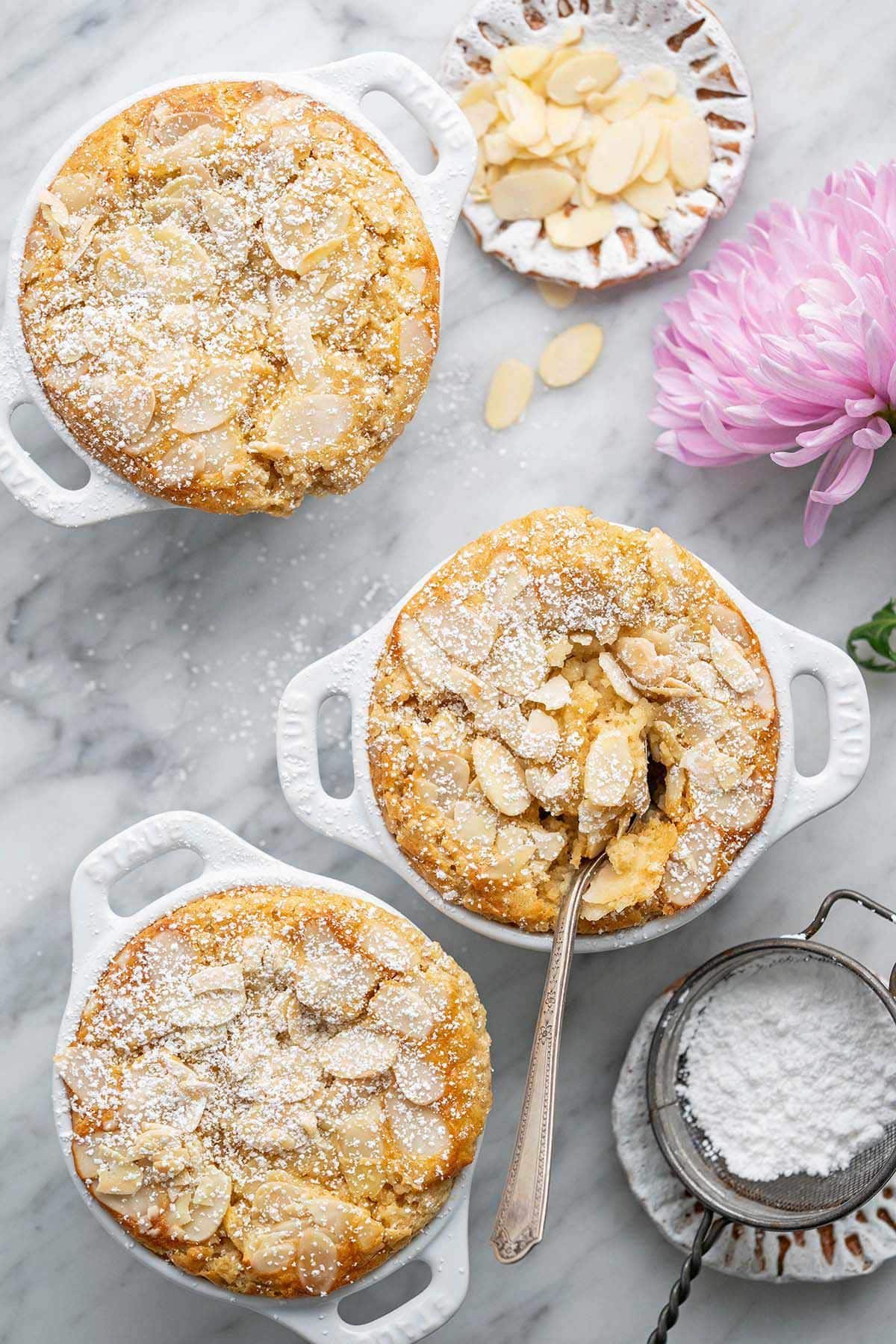 Three individual white ramekins of baked oatmeal topped with sliced almonds and powdered sugar, on a marble surface with a pink flower