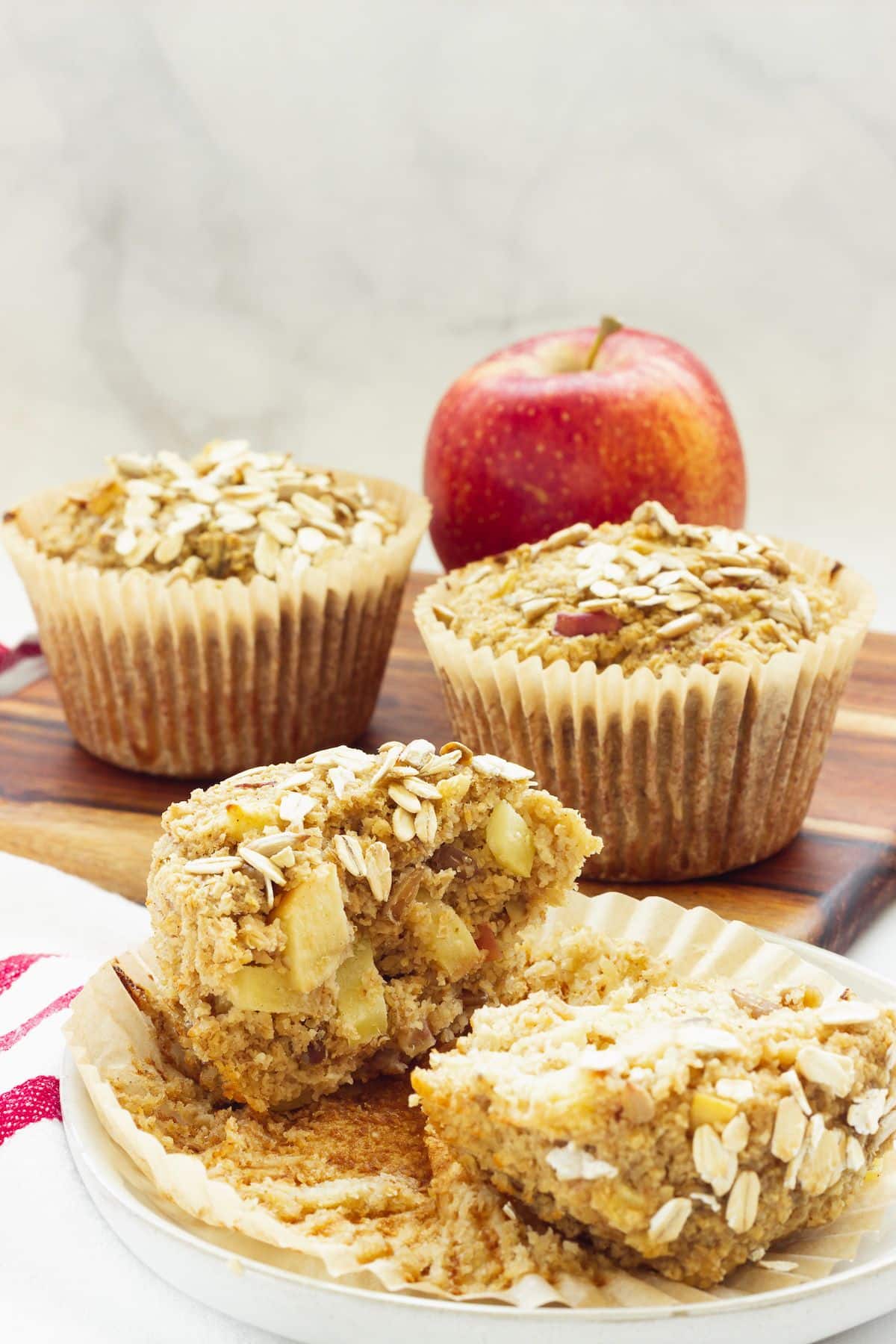 Three apple bran muffins with oat topping on a wooden cutting board, one split open to show a moist interior, with a red apple in the background"