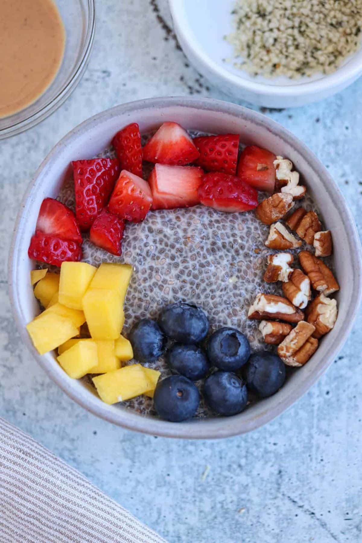 Overhead view of a chia pudding bowl topped with fresh strawberries, raspberries, blueberries, mango chunks, pecans, and hemp seeds