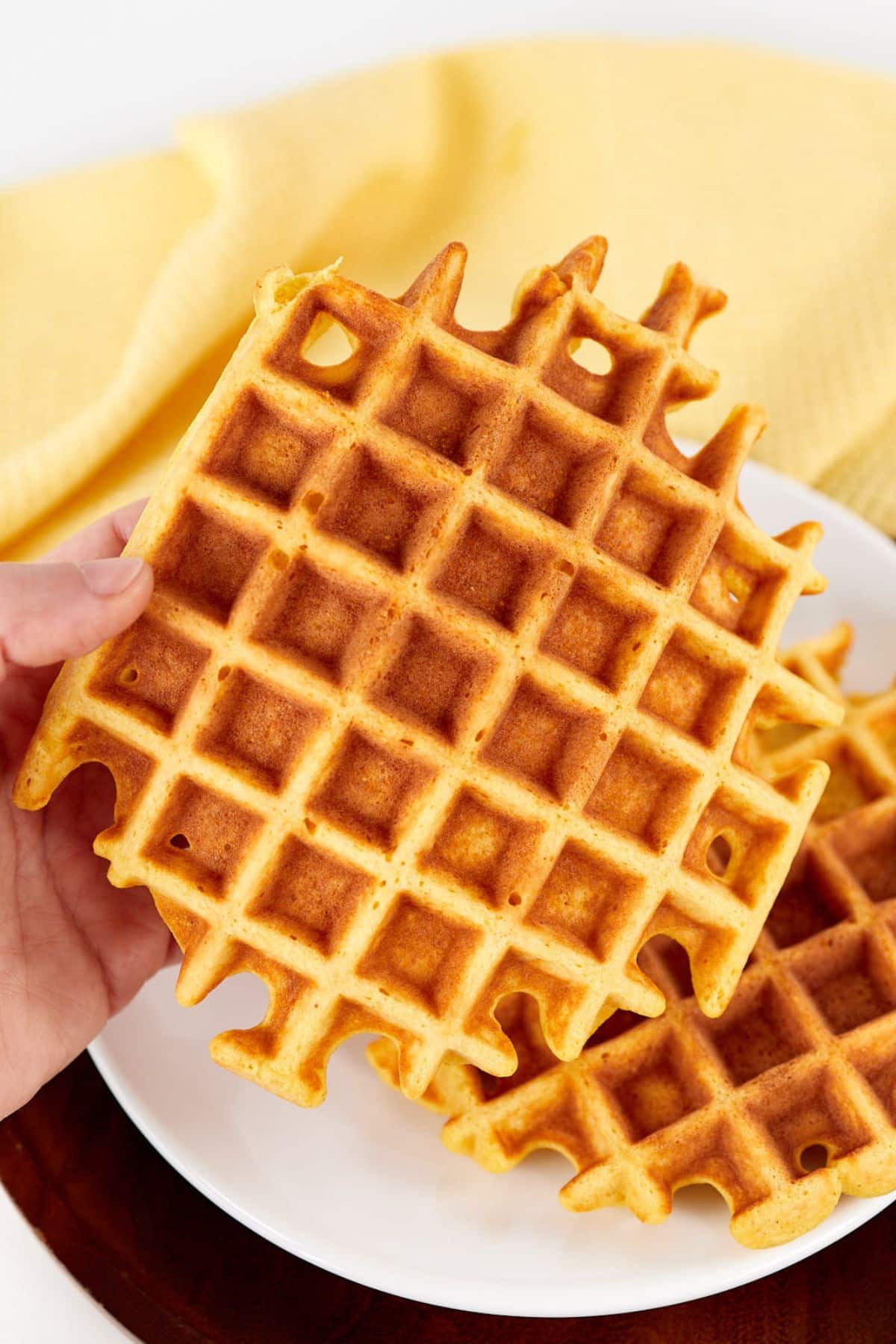 Hand holding a large golden crispy waffle over a white plate with a yellow cloth in the background.
