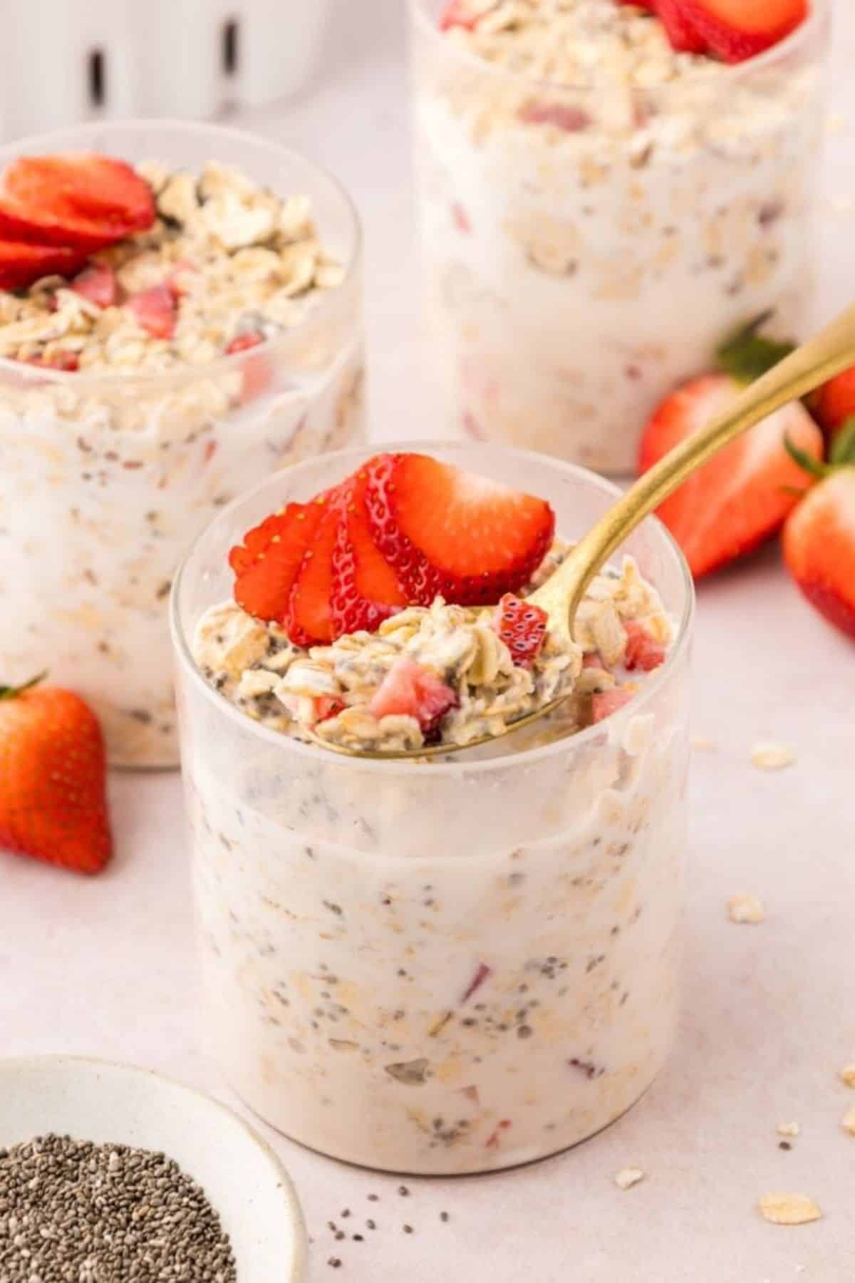 Three jars of strawberry overnight oats with chia seeds and fresh strawberries on a light pink surface.