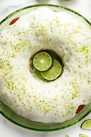 Square overhead crop of key lime pound cake with cream cheese glaze, lime zest, and sliced limes on green plate.