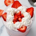 Overhead close-up of a Ninja Creami pint cup filled with strawberry protein ice cream, topped with piped whipped cream swirls and chopped and sliced fresh strawberries, on a white marble surface.