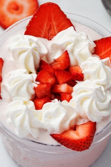 Overhead close-up of a Ninja Creami pint cup filled with strawberry protein ice cream, topped with piped whipped cream swirls and chopped and sliced fresh strawberries, on a white marble surface.