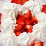 Wide overhead shot of Ninja Creami strawberries and cream protein ice cream topped with whipped cream rosettes and fresh sliced and chopped strawberries in a clear pint cup on a white marble surface.