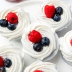 Six no-bake mini blueberry cheesecakes on a white plate, each topped with whipped cream, blueberries, and a raspberry.