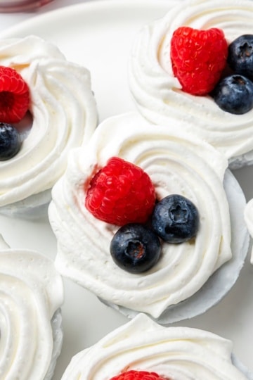 Six no-bake mini blueberry cheesecakes on a white plate, each topped with whipped cream, blueberries, and a raspberry.