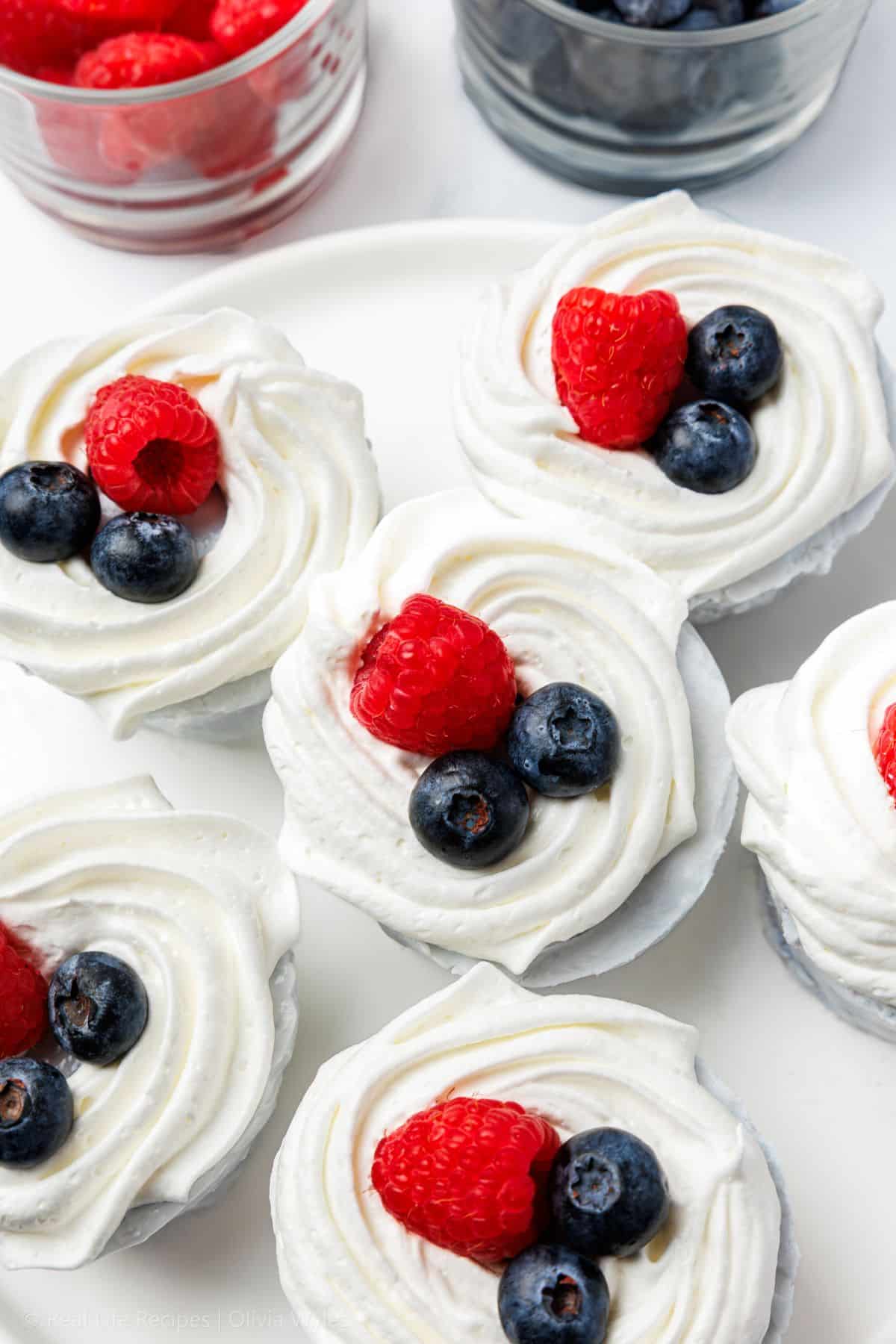 Six no-bake mini blueberry cheesecakes arranged on a white plate, each finished with whipped cream, blueberries, and raspberries.
