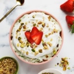 Overhead view of pistachio fluff in a pink ceramic bowl with strawberry slices, pistachios, and whipped cream garnish, surrounded by ingredient bowls on marble surface.