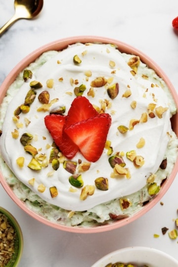 Overhead view of pistachio fluff in a pink ceramic bowl with strawberry slices, pistachios, and whipped cream garnish, surrounded by ingredient bowls on marble surface.