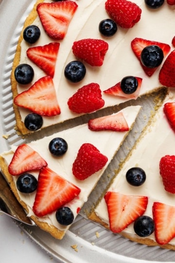 Red white and blue fruit pizza with strawberries, raspberries, and blueberries on a cream cheese frosting crust with a slice removed.