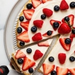 Overhead view of a sliced red white and blue fruit pizza topped with fresh strawberries, raspberries, and blueberries on a white plate.