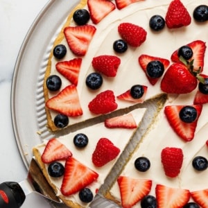 Overhead view of a sliced red white and blue fruit pizza topped with fresh strawberries, raspberries, and blueberries on a white plate.