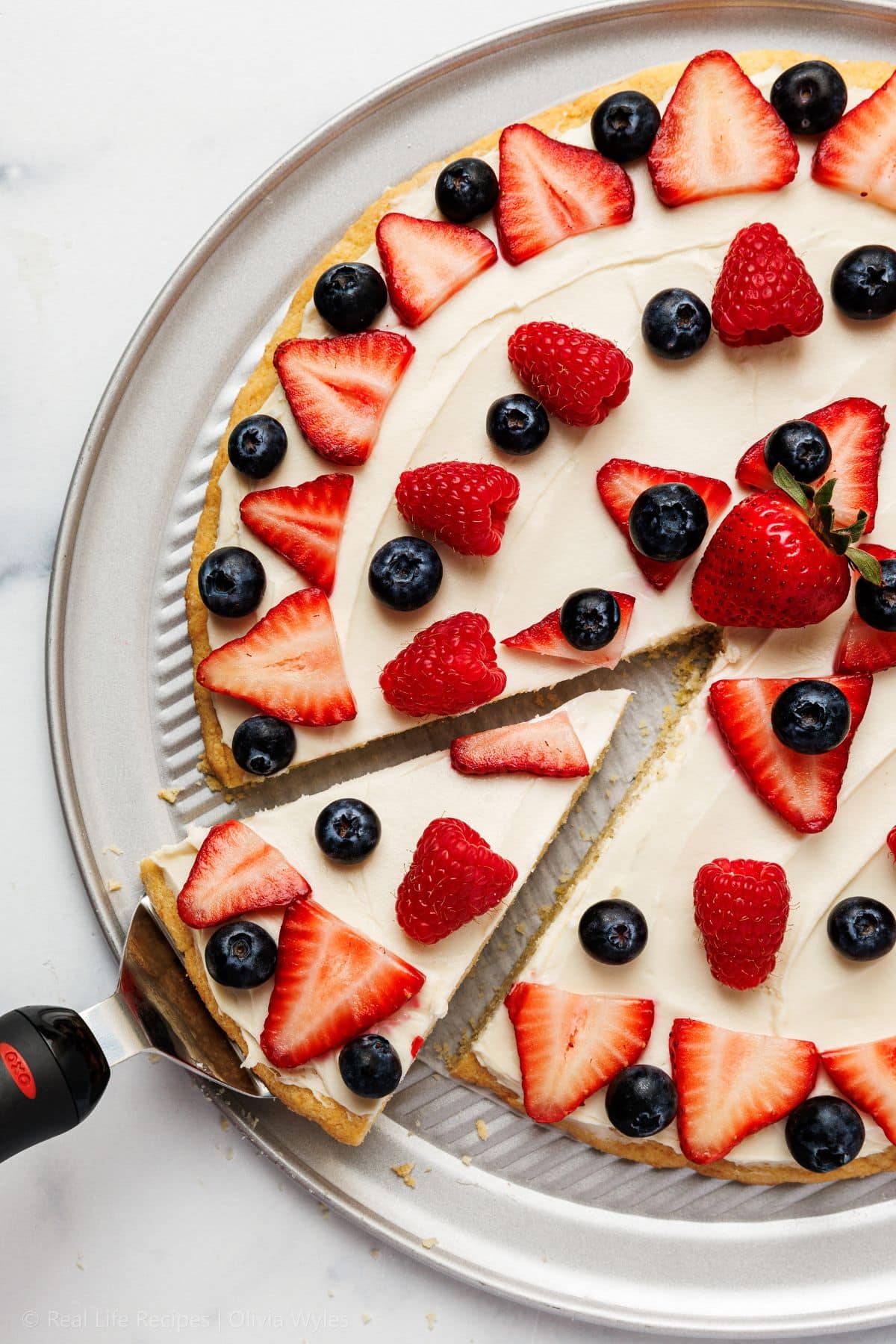 Overhead view of a sliced red white and blue fruit pizza topped with fresh strawberries, raspberries, and blueberries on a white plate.