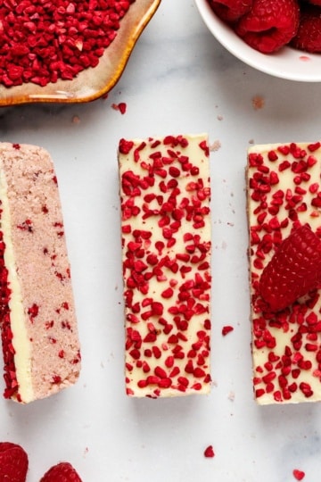 white chocolate raspberry protein bars topped with freeze-dried raspberries on a marble surface.Five white chocolate raspberry protein bars topped with freeze-dried raspberries on a marble surface.