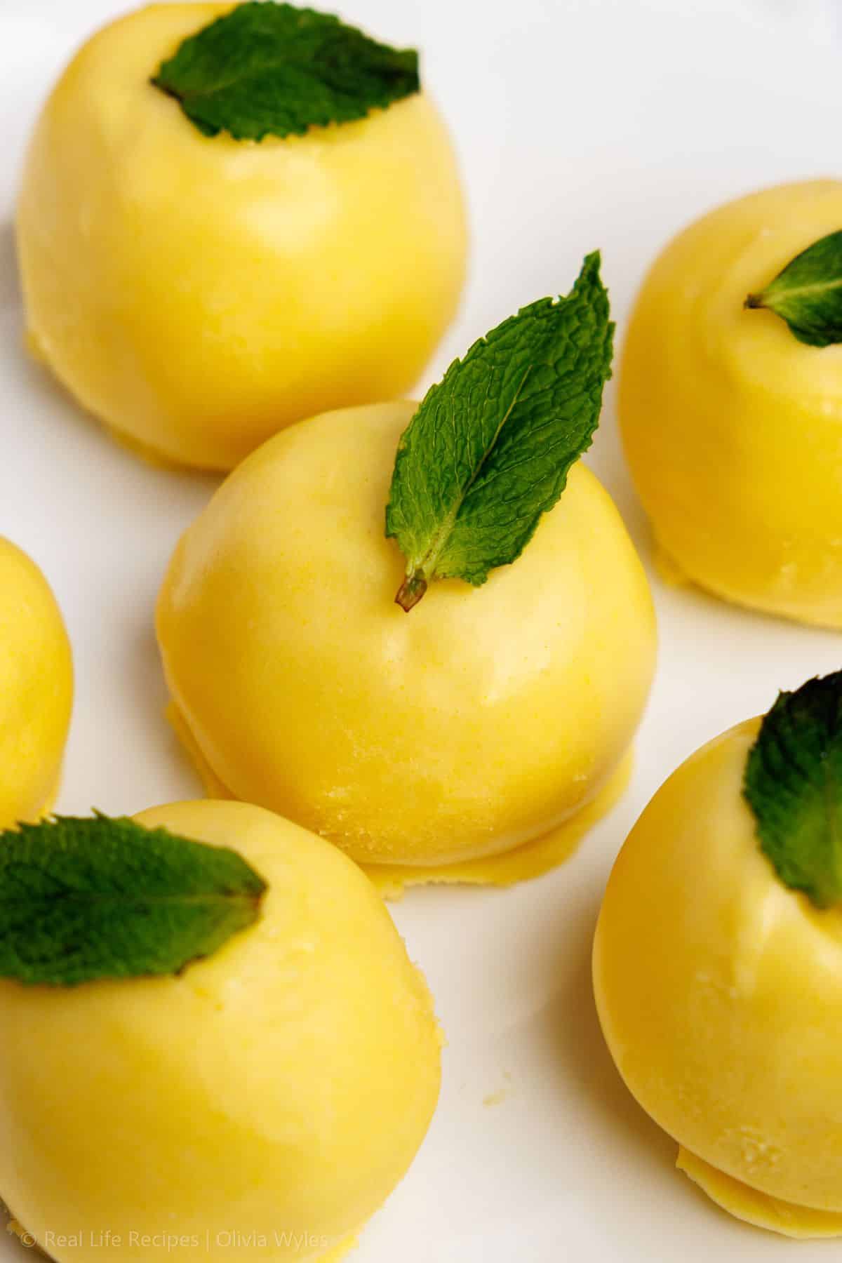 Overhead view of lemon truffles topped with fresh mint leaves on a white plate, whole lemons nearby.