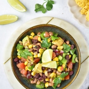 Overhead view of Cowboy Caviar bean salad in a bowl with lime wedges, cilantro, and tortilla chips nearby.