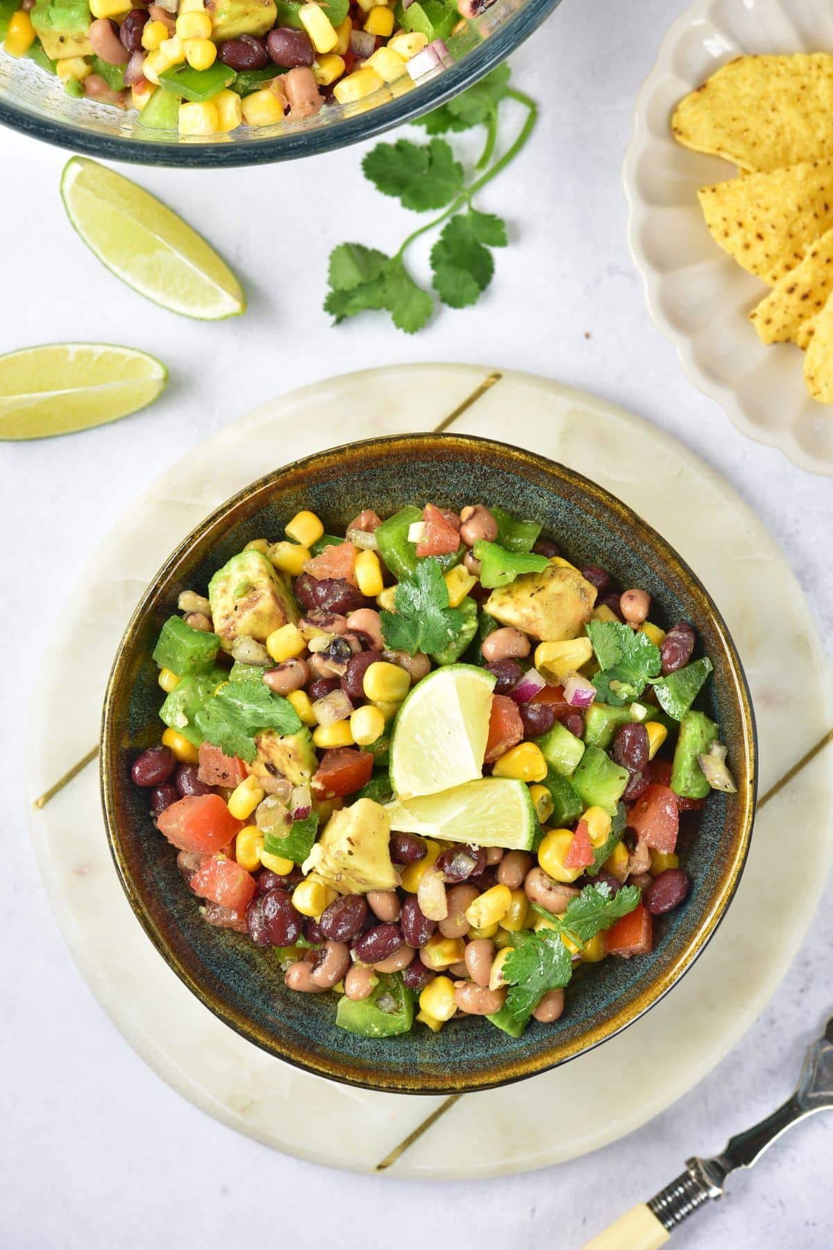 Overhead view of Cowboy Caviar bean salad in a bowl with lime wedges, cilantro, and tortilla chips nearby.