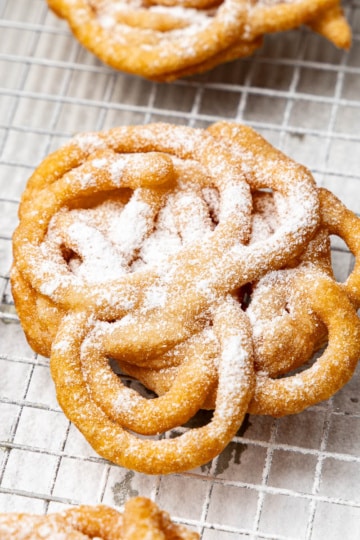Close-up of a mini funnel cake dusted with powdered sugar on a wire rack, with more cakes blurred behind it.