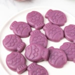 Purple Sugar-Free Gummy Fish stacked on a white plate, with one red fish gummy in a glass bowl behind