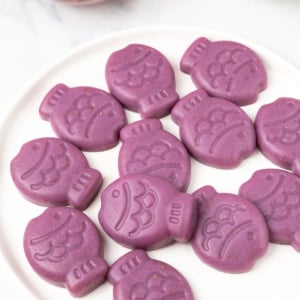 Purple Sugar-Free Gummy Fish stacked on a white plate, with one red fish gummy in a glass bowl behind