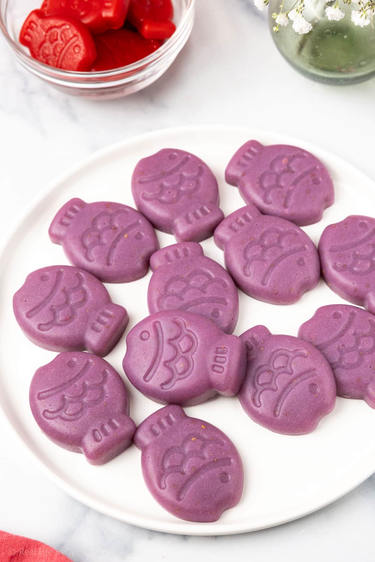 Purple Sugar-Free Gummy Fish stacked on a white plate, with one red fish gummy in a glass bowl behind